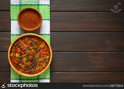Vegan goulash made of soy meat (textured vegetable protein), capsicum, tomato and onion in wooden bowl, paprika powder in small bowl, photographed overhead on dark wood with natural light