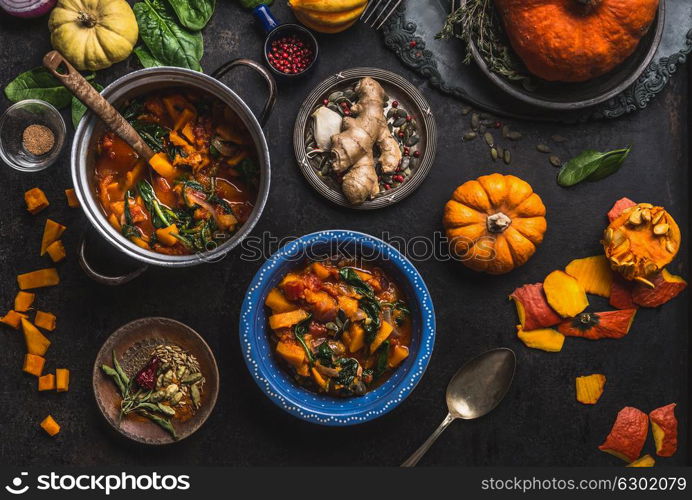 Vegan pumpkin stew dish with spinach served in bowl with spoon on dark kitchen table background with pot and ingredients. Healthy seasonal food and clean eating concept