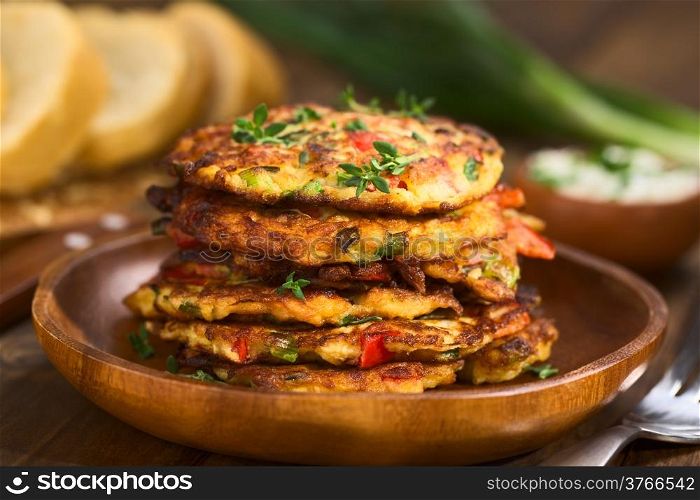 Vegetable and egg fritter made of zucchini, red bell pepper, eggs, green onions and thyme piled on a wooden plate with baguette slices and yogurt dip in the back (Selective Focus, Focus on the front of the thyme sprig on the top of the fritters and on the front of the top fritters)