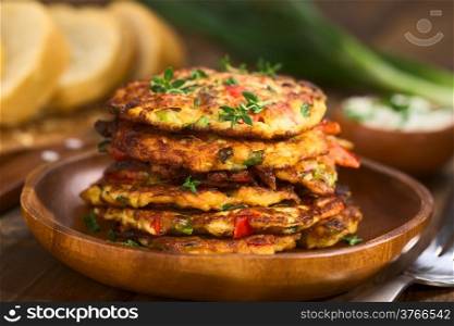 Vegetable and egg fritter made of zucchini, red bell pepper, eggs, green onions and thyme piled on a wooden plate with baguette slices and yogurt dip in the back (Selective Focus, Focus on the front of the thyme sprig on the top of the fritters and on the front of the top fritters)