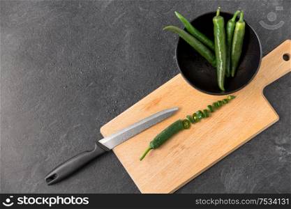 vegetable, food and culinary concept - close up of green chili peppers and kitchen knife on wooden cutting board on slate stone background. green chili peppers and knife on cutting board