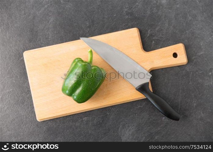 vegetable, food and culinary concept - close up of green pepper and kitchen knife on wooden cutting board on slate stone background. green pepper and kitchen knife on cutting board