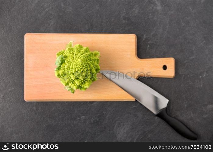 vegetable, food and culinary concept - close up of romanesco broccoli and kitchen knife on wooden cutting board on slate stone background. romanesco broccoli and knife on cutting board