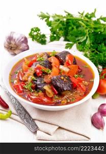 Vegetable ragout with eggplant, tomatoes, bell peppers, onions and spices in a plate on napkin, garlic, parsley, hot peppers and a fork on the background of light wooden board