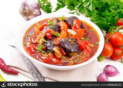 Vegetable ragout with eggplant, tomatoes, bell peppers, onions and spices in a plate on towel, garlic, parsley, hot peppers and a fork on the background of light wooden board