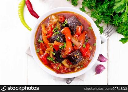 Vegetable ragout with eggplant, tomatoes, bell peppers, onions and spices in a plate on napkin, garlic, parsley, hot peppers and a fork on wooden board background from above