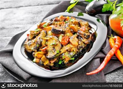 Vegetable ragout with eggplant, tomatoes, sweet and hot peppers, onions, carrots, fried with herbs and spices in a plate on a towel, garlic, parsley on wooden board background