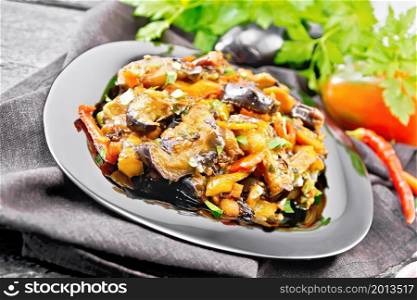 Vegetable ragout with eggplant, tomatoes, sweet and hot peppers, onions, carrots, fried with herbs and spices in a plate on a towel, garlic, parsley on dark wooden board background