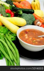 Vegetable soup in a bowl surrounded by fresh vegetables in cluding squash, carrot, green beans, mushrooms, celery and bell peppers. Vegetable Soup