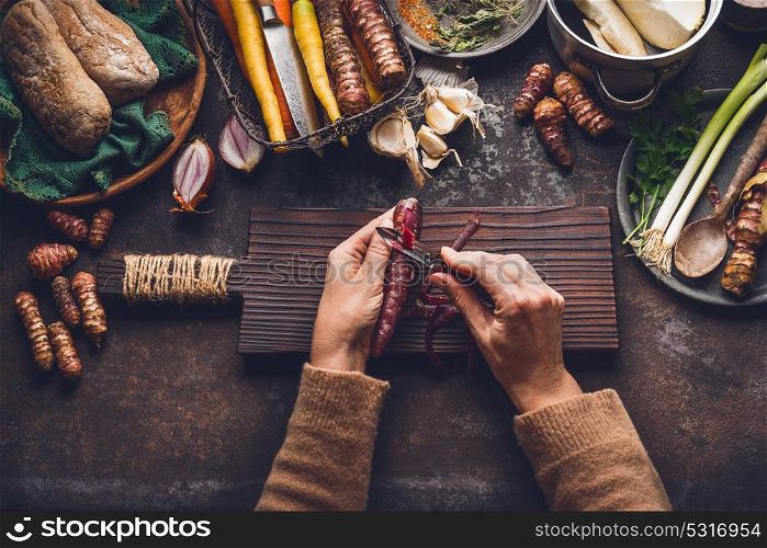 Vegetables cooking and eating. Women female hand peeling carrots on dark rustic kitchen table background with various root vegetables ingredients, top view. Healthy clean eating or diet food concept