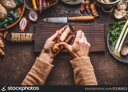 Vegetables cooking and eating. Women female hand peeling Jerusalem artichokes on dark rustic kitchen table background with various root vegetables ingredients, top view. Healthy eating or diet food.