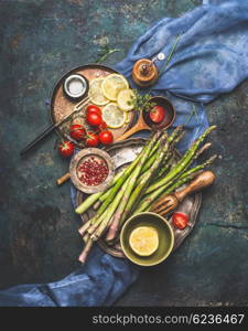 Vegetables cooking ingredients: green asparagus,tomatoes, lemon and flavoring on dark rustic background , top view