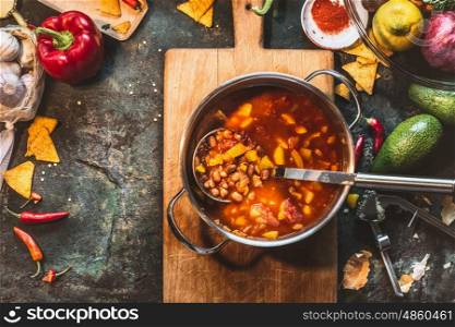 Vegetarian Bean soup in cooking pot with ladle on wooden cutting board with ingredients on dark rustic background, top view, frame. Mexican cuisine