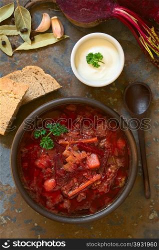 Vegetarian Borscht soup of Ukrainian origin made of beetroot, carrot, cabbage, potato, onion and celery in rustic bowl, wholegrain bread, sour cream, ingredients on the side, photographed overhead with natural light (Selective Focus, Focus on the soup)