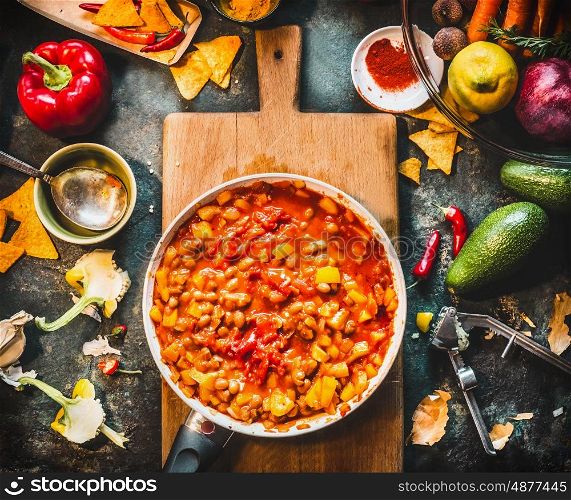Vegetarian chili con carne dish in pan on wooden cutting board with spices and vegetables cooking ingredients on dark kitchen table background, top view. Mexican cuisine