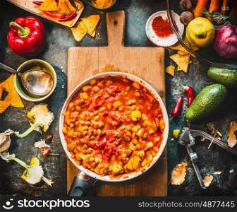 Vegetarian chili con carne dish in pan on wooden cutting board with spices and vegetables cooking ingredients on dark kitchen table background, top view. Mexican cuisine