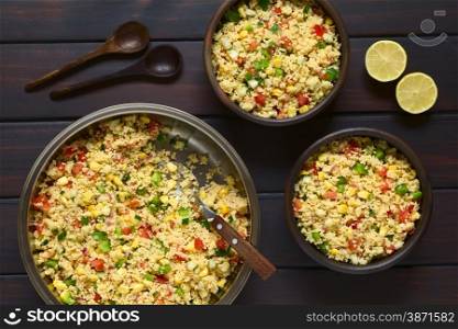 Vegetarian couscous salad made with bell pepper, tomato, cucumber, red onion and sweet corn kernels, wooden spoons and lemon on the side. Photographed overhead on dark wood with natural light.