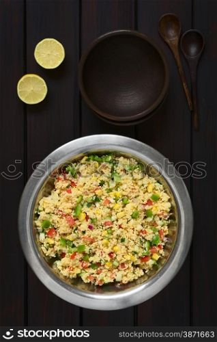 Vegetarian couscous salad made with bell pepper, tomato, cucumber, red onion and sweet corn kernels, served in salad bowl with rustic bowls, wooden spoons and lemon on the side. Photographed overhead on dark wood with natural light.