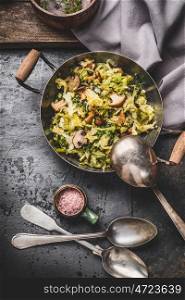 Vegetarian dish with cabbage and mushrooms in rustic cooking pot with spoons on dark kitchen table, top view, place for text