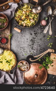 Vegetarian dishes with yellow rice, cabbage and mushrooms in cooking pots on dark rustic country kitchen table with spoons, top view, frame