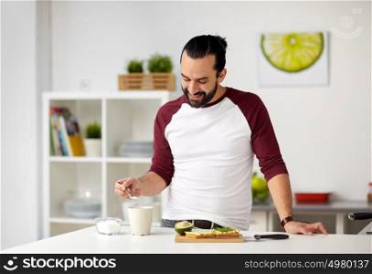 vegetarian food, healthy eating, people and diet concept - man adding sugar to tea or coffee cup and having vegetable sandwiches for breakfast at home kitchen. man adding sugar to cup for breakfast at home