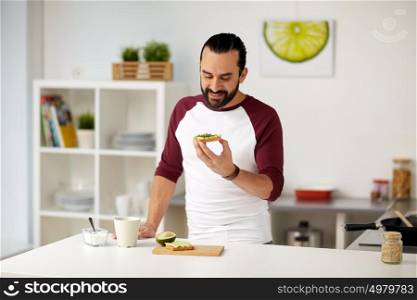 vegetarian food, healthy eating, people and diet concept - man having avocado sandwiches for breakfast at home kitchen. man eating avocado sandwiches at home kitchen