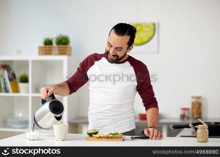 vegetarian food, healthy eating, people and diet concept - man with kettle making tea and having vegetable sandwiches for breakfast at home kitchen. man with kettle making tea for breakfast at home