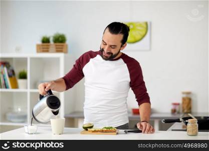 vegetarian food, healthy eating, people and diet concept - man with kettle making tea and having vegetable sandwiches for breakfast at home kitchen. man with kettle making tea for breakfast at home