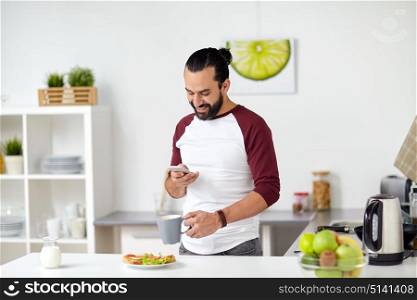 vegetarian food, healthy eating, people, technology and breakfast concept - man with smartphone, coffee and vegetable sandwiches at home kitchen. man with smartphone having breakfast at home