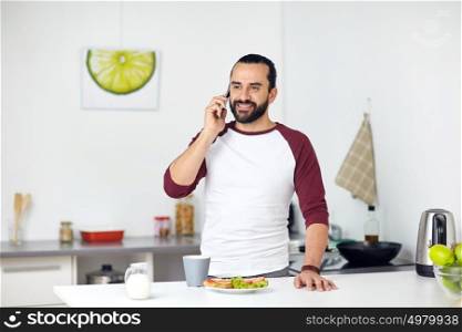 vegetarian food, healthy eating, people, technology and diet concept - man having vegetable sandwiches with coffee for breakfast and calling on smartphone at home kitchen. man calling on smartphone and eating at home