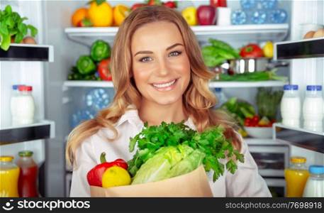 Vegetarian lifestyle, portrait of a beautiful woman standing near open fridge full of different fresh vegetables, organic nutrition, weight control, healthy eating concept 