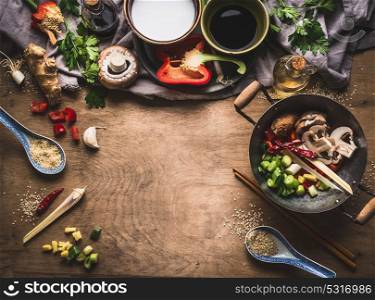 Vegetarian stir fry cooking preparation on wooden background with various vegetables, wok, coconut milk, seeds and kitchen utensils, top view, frame. Asian cuisine. Healthy eating and food concept