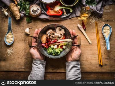 Vegetarian stir fry cooking preparation. Women female hands holding little wok pot with chopped vegetables for stir fry on kitchen table background with vegetables ingredients, top view