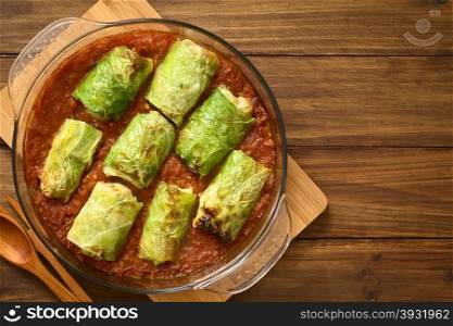 Vegetarian stuffed savoy cabbage rolls filled with wholegrain rice, pepper, onion and carrot, baked on tomato sauce in pan, photographed overhead on dark wood with natural light