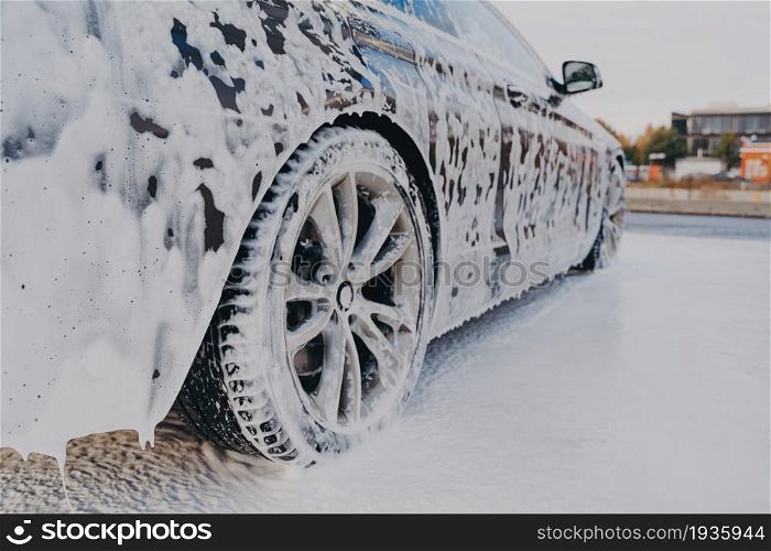 Vehicle covered in white soapy foam during regular carwash outdoors, auto getting professional wash with soap at washing center, cleaning car exterior surface at self-service station. Vehicle in white soapy foam during regular car wash outdoors, auto getting wash with soap