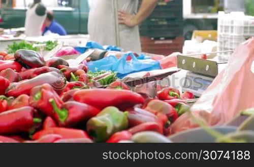 Vendors selling red sweet pepper on street market. Woman paying for vegetables, getting change and taking the packet. Seller adding peppers on the stall