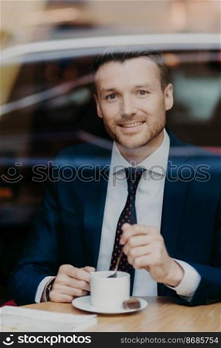 Verrtical shot of delighted handsome male professional worker in black suit, drinks aromatic beverage, looks joyfully at camera, poses in restaurant, sits near window, has coffee break after work
