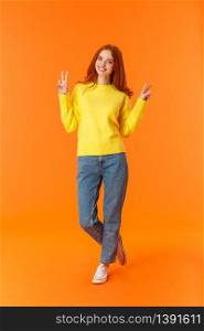Vertical full-length cute lovely redhead teenage girl in jeans and winter yellow sweater standing with peace signs over orange background, smiling posing express carefree joy emotions.. Vertical full-length cute lovely redhead teenage girl in jeans and winter yellow sweater standing with peace signs over orange background, smiling posing express carefree joy emotions