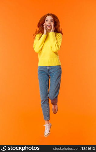 Vertical full-length portrait pretty redhead caucasian girl in casual jeans, yellow sweater, look surprised and excited, holding hands near face impressed, jumping over orange background happy.. Vertical full-length portrait pretty redhead caucasian girl in casual jeans, yellow sweater, look surprised and excited, holding hands near face impressed, jumping over orange background happy