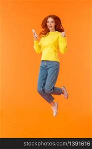 Vertical full-length shot carefree, excited and pleased good-looking ginger girl in sweater, jeans, jumping over orange background, holding smartphone show thumbs-up and smiling approvingly.. Vertical full-length shot carefree, excited and pleased good-looking ginger girl in sweater, jeans, jumping over orange background, holding smartphone show thumbs-up and smiling approvingly