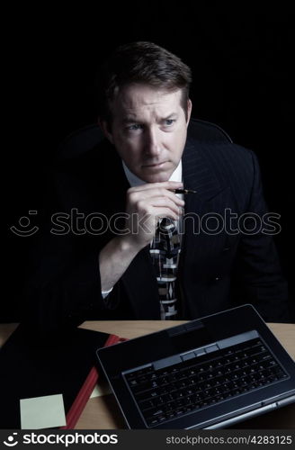 Vertical image of business man, looking at computer screen while holding pen, working late with black background