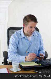 Vertical image of mature man looking at laptop, with pencil and cell phone in hand, while working. White walls in background.