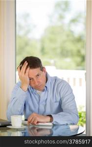 Vertical image of mature man showing depression by holding his head in one hand while working from home with bright daylight coming in from window in background