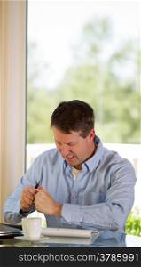 Vertical image of mature man showing stress by crumbling paper while working from home with bright daylight coming in from window in background
