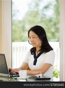 Vertical image of mature woman relaxed during typing on laptop while working from home with blurred out daylight coming in from window in background