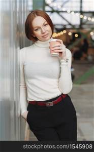 Vertical image of thoughtful young woman wears white jumper and black trousers, holds takeaway coffee, dreams about something pleasant, poses outdoor, keeps hand in pocket. People and lifestyle