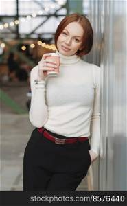 Vertical image of thoughtful young woman wears white jumper and black trousers, holds takeaway coffee, dreams about something pleasant, poses outdoor, keeps hand in pocket. People and lifestyle