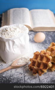 Vertical image with a bag of flour, a wooden spoon, egg, Belgian waffles and an open book, on a vintage black wooden background. A baking concept.