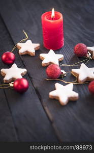 Vertical image with a red lit candle on a black wooden table, surrounded by stars shape cookies and red globes, for a Christmas atmosphere.