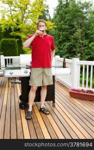 Vertical photo of mature man drinking a beer out of a glass with barbecue cooker and seasonal trees in background